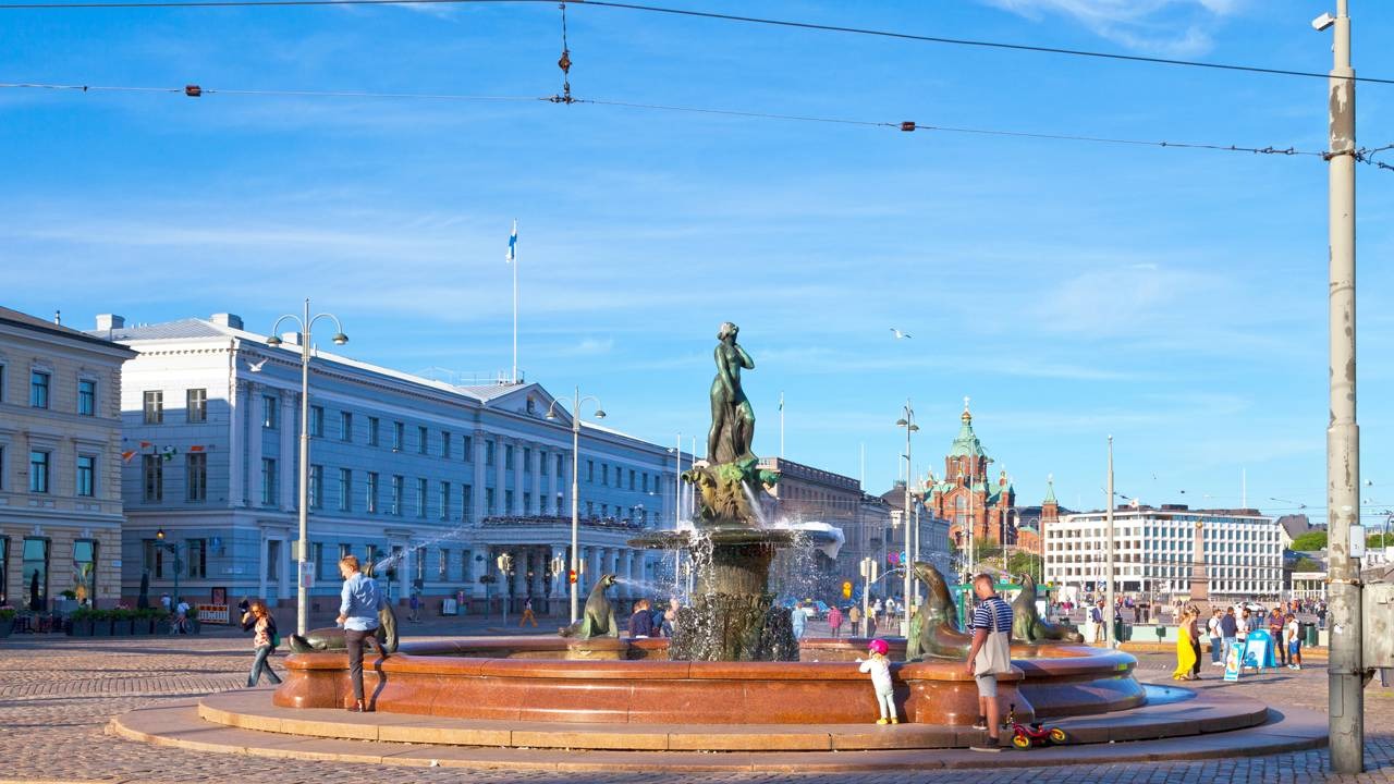A praça Havis Amanda, em Helsinque, ilustra o cenário urbano e a qualidade de vida que contribuem para a Finlândia ser o país mais feliz do mundo. Créditos: depositphotos.com / BreizhAtao Fonte Havis Amanda em praça ensolarada de Helsinque com pessoas, prédios e bandeira da Finlândia.
