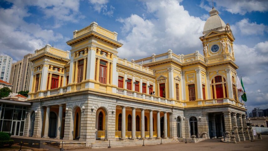O Palácio da Liberdade, com sua arquitetura imponente, evoca os cenários palacianos da série 'Bridgerton' em Minas Gerais. Palácio amarelo neoclássico com torre do relógio, colunas, janelas vermelhas e bandeiras sob céu azul.