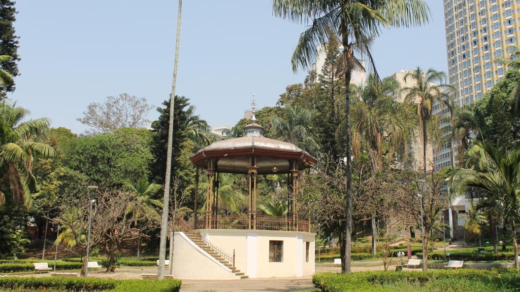 O Parque Municipal Américo Renné Giannetti, com seu coreto, remete aos cenários de socialização da série 'Bridgerton'. Coreto octogonal bege e marrom com escadaria em parque, cercado por palmeiras e edifícios altos.