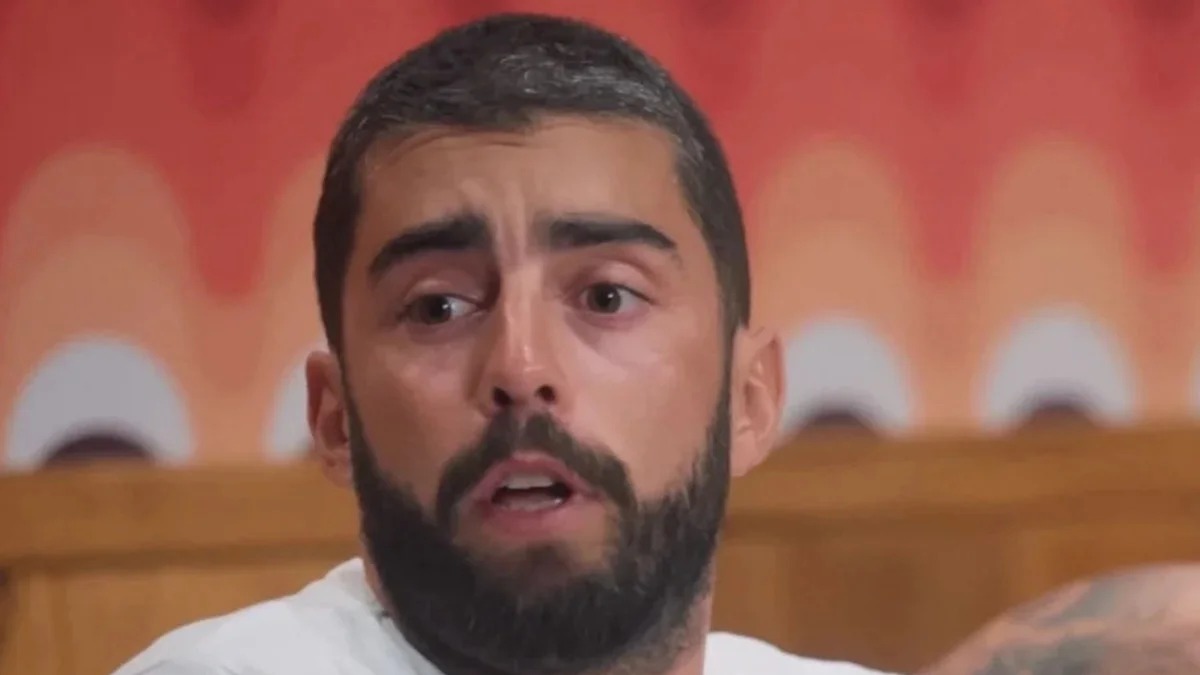 Pedro Scooby reage a boatos sobre sua vida pessoal e nega affair após separação de Cintia Dicker. Foto: YouTube/Reprodução Close-up de homem barbudo, boca entreaberta, olhando para o lado, camisa branca