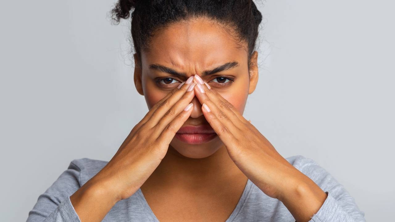 O desconforto nasal é um dos sintomas da rinite, intensificado com o tempo seco e a baixa umidade. Mulher com as mãos sobre o nariz, testa franzida, olhos abertos, em fundo cinza