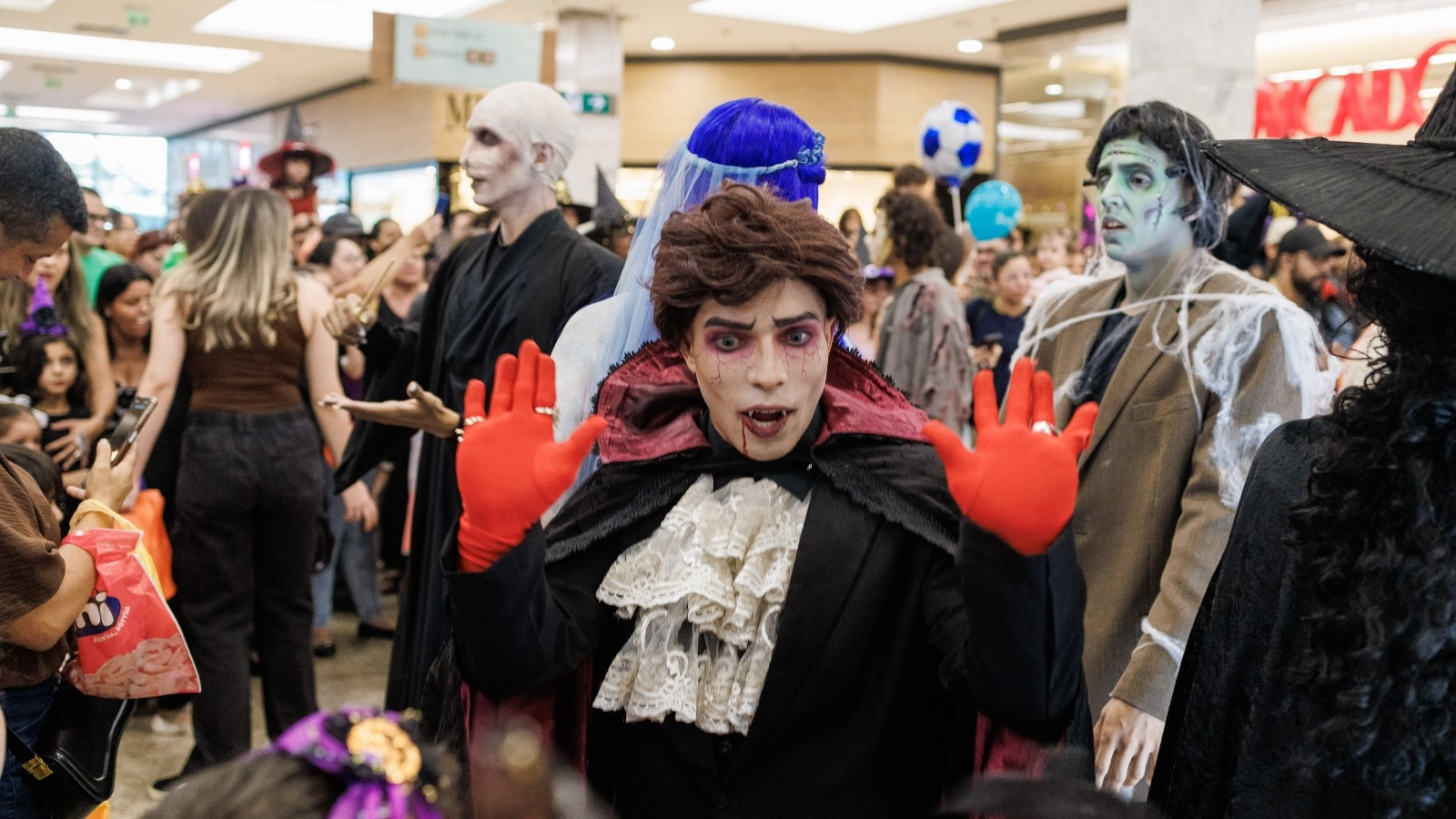 Pessoa vestida de vampiro com capa, maquiagem e luvas vermelhas posa entre outros fantasiados em shopping