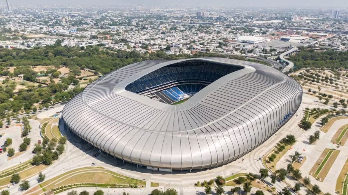 Estádio BBVA, em Monterrey Vista aérea de estádio moderno, exterior prateado, arquibancadas azuis, rodeado por vegetação e cidade.