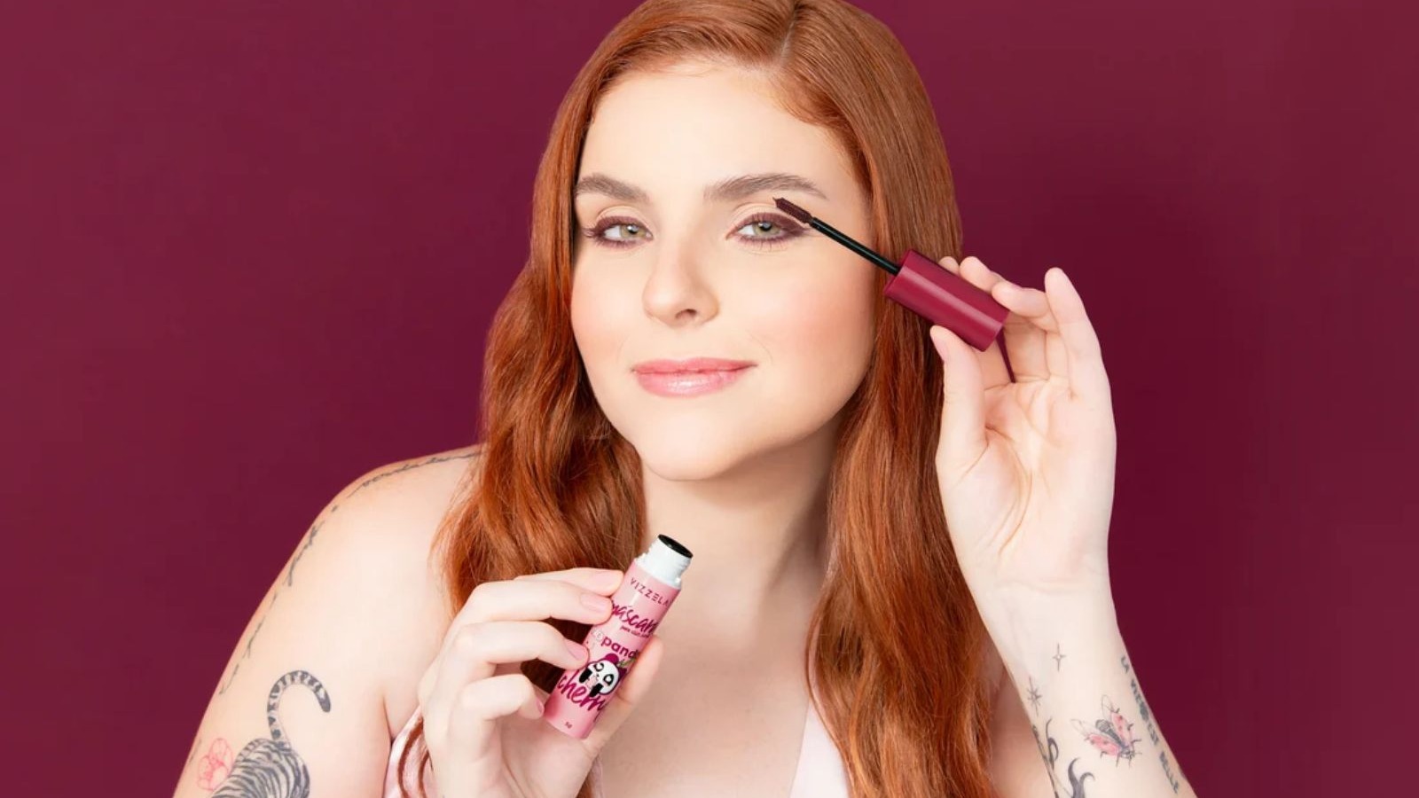 Mulher ruiva aplica m&aacute;scara de c&iacute;lios bord&ocirc; Vizzela em olho, segurando outro produto pink