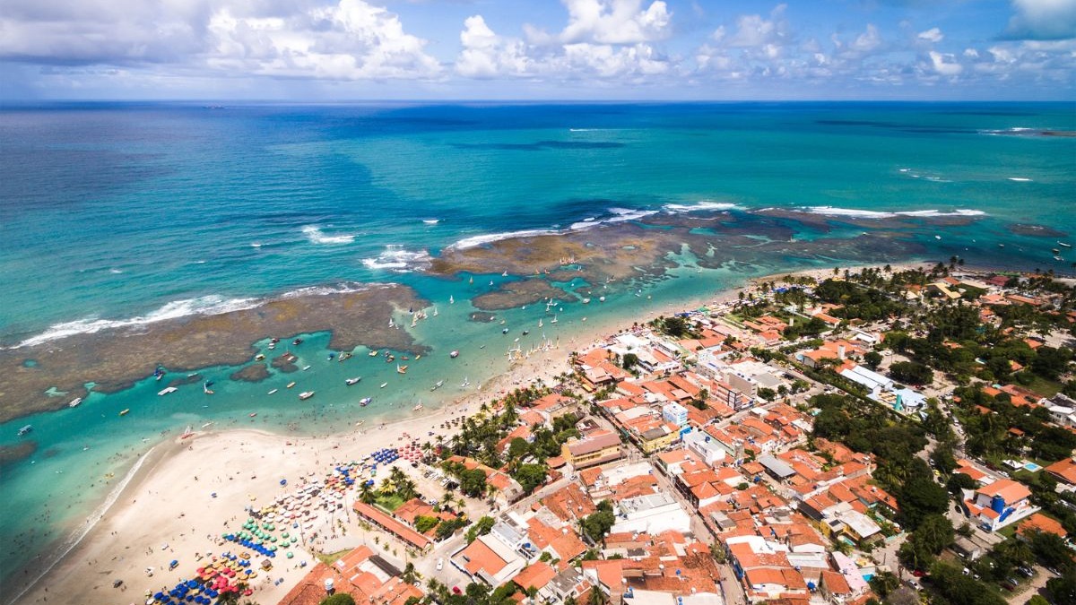 Vista aérea de praia movimentada com mar azul, jangadas, recifes, guarda-sóis e cidade.