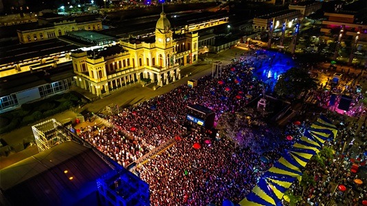 Vista aérea de multidão em evento noturno na Praça da Estação, com prédio histórico iluminado e projeções coloridas