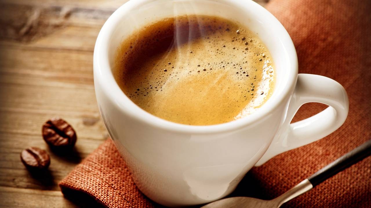 Descubra como uma pitada de sal pode surpreender, neutralizando o amargor e realçando o sabor do café, conforme truques culinários. Xícara branca de café fumegante com grãos e colher sobre mesa de madeira