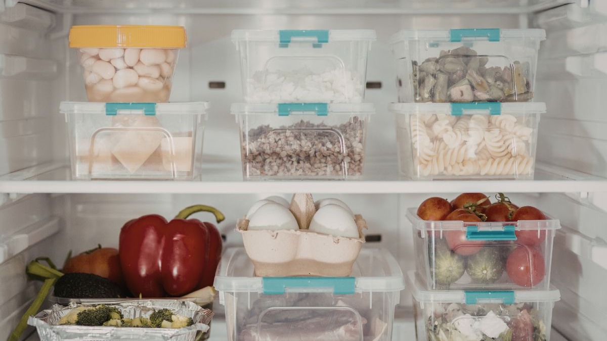 Geladeira aberta com prateleiras cheias de alimentos organizados em potes pl&aacute;sticos transparentes.