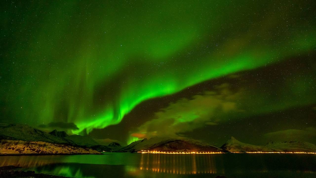 A Noruega, conhecida por seus fenômenos naturais, oferece espetáculos como a Aurora Boreal em suas noites invernais. Aurora Boreal verde vibrante no céu noturno, com montanhas, lago e luzes de uma cidade.