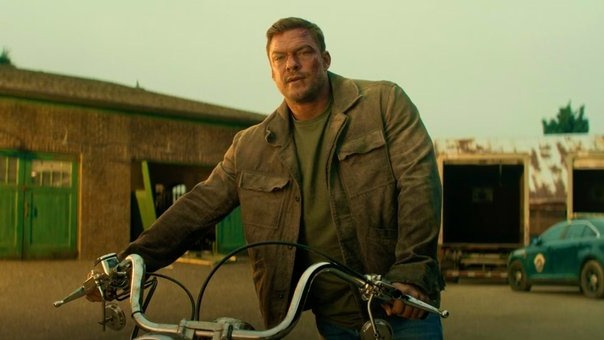 Alan Ritchson, o protagonista de Reacher, promete uma temporada com nível de ação sem precedentes. Alan Ritchson, ator, em jaqueta, camiseta verde, inclinado sobre motocicleta.