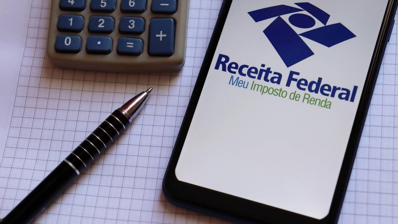Contribuintes podem verificar os valores da restituição automática do IRPF pelo aplicativo da Receita Federal. Celular mostra 'Receita Federal Meu Imposto de Renda', calculadora e caneta em bloco de anotações