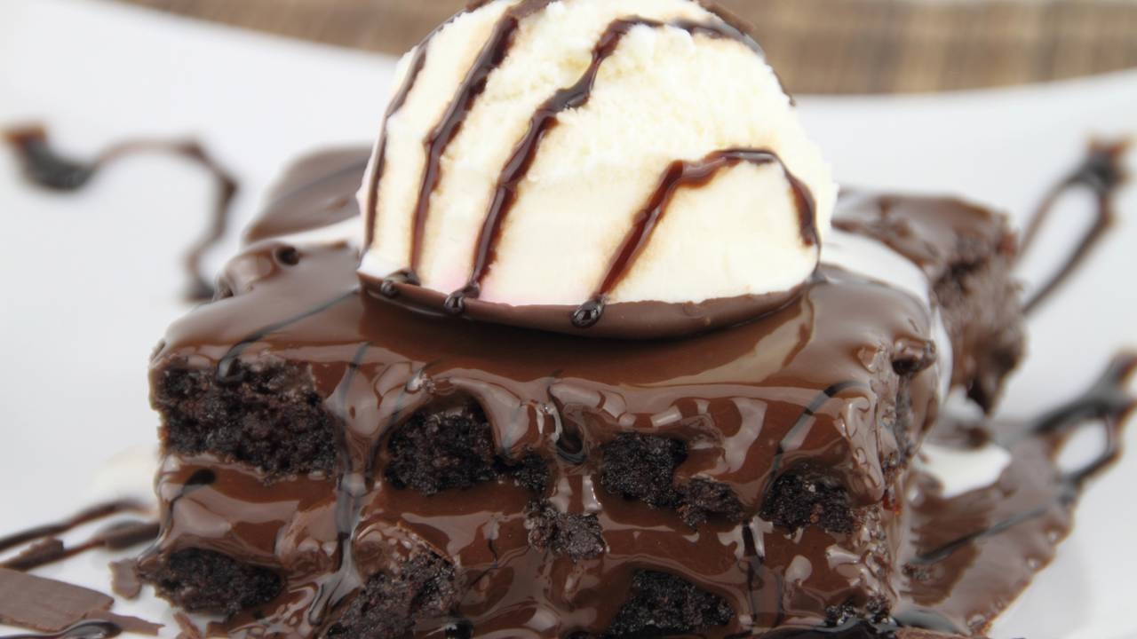 Aprenda a preparar um brownie com textura macia e casquinha crocante, ideal para servir com sorvete e calda. Pedaço de brownie de chocolate coberto com calda e sorvete de baunilha