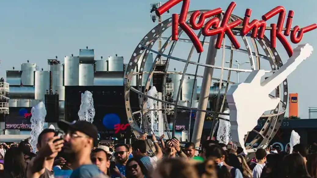 Multidão em frente ao letreiro Rock in Rio com globo, guitarra, e fontes sob céu azul, placa Itaú