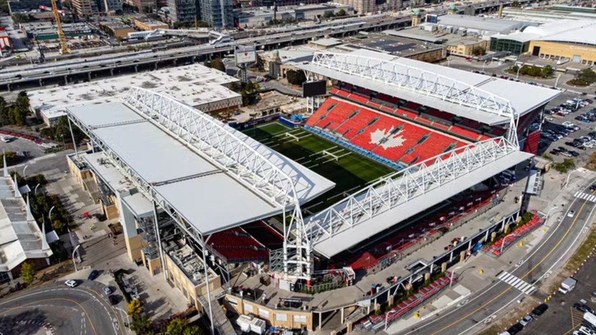 O BMO Field em Toronto, com sua arquibancada exibindo a folha de bordo do Canadá, será um dos palcos da Copa do Mundo de 2026. Vista aérea de estádio com campo verde, assentos vermelhos formando folha de bordo, tetos brancos.