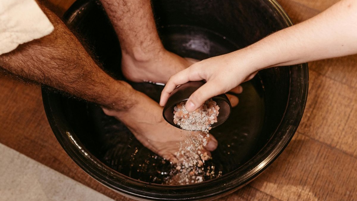 A receita caseira de escalda-pés, com sal e vinagre, pode ser uma solução simples para aliviar dores e odores nos membros inferiores Mão despeja sal rosa e branco de tigela preta em bacia com pés na água