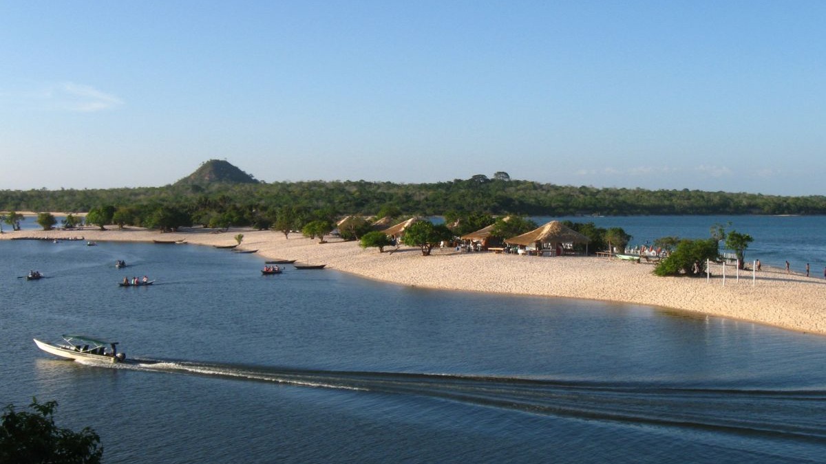 Vista a&eacute;rea de rio com praia de areia branca, barcos, quiosques, vegeta&ccedil;&atilde;o e colina.