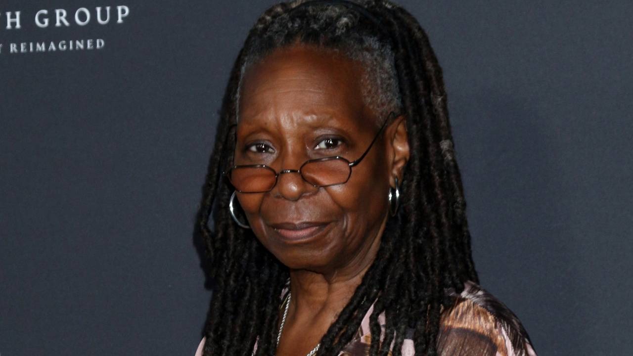 Whoopi Goldberg, que viu sua estatueta do Oscar ir parar no lixo e ser encontrada por um segurança. Whoopi Goldberg com dreadlocks e óculos sorrindo levemente em fundo escuro com texto desfocado
