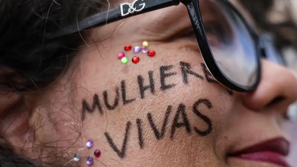 O Levante Mulheres Vivas realizando ato na área central de Brasília para denunciar o feminicídio. Rosto de mulher, óculos, 'MULHERES VIVAS' na bochecha com strass coloridos