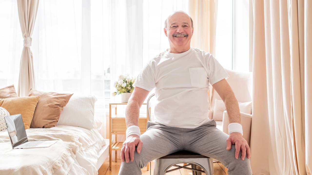 Homem idoso sorridente de camiseta branca sentado em cadeira metálica em quarto iluminado