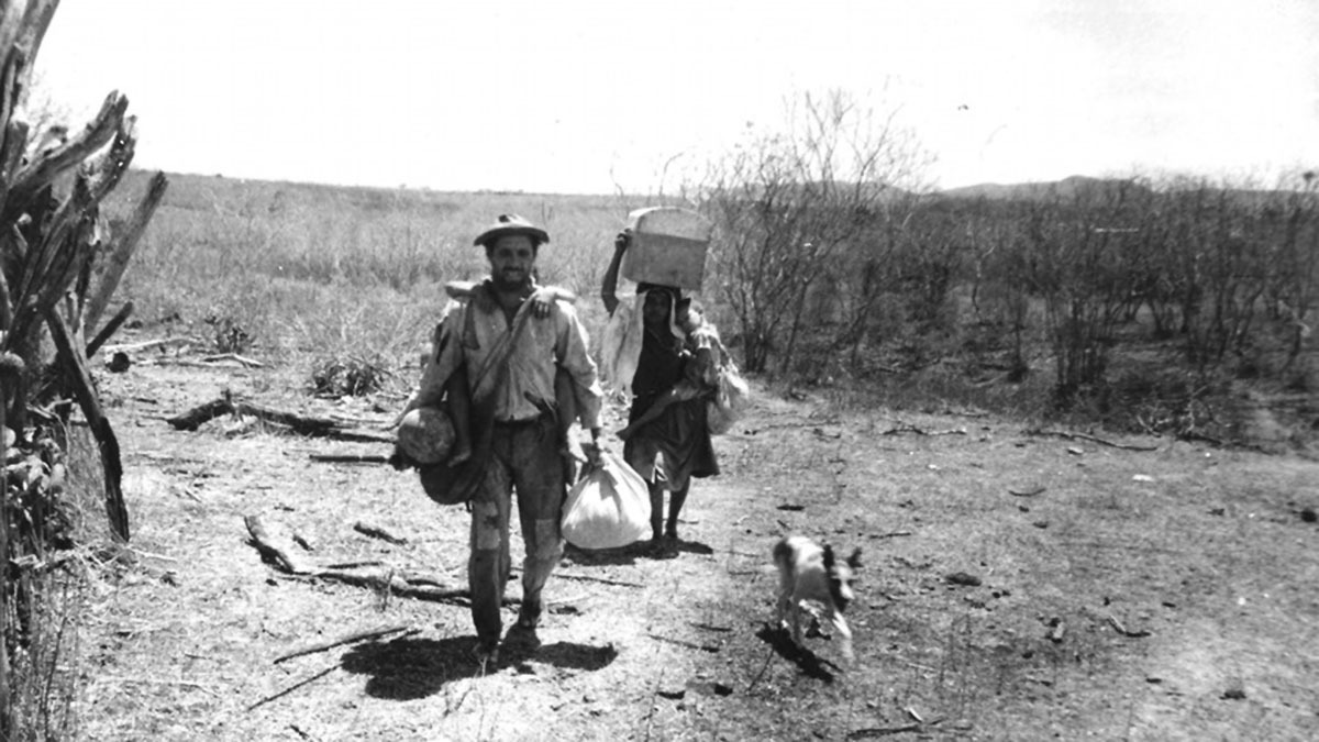 Foto em preto e branco de duas pessoas e um cachorro caminhando em estrada de terra árida