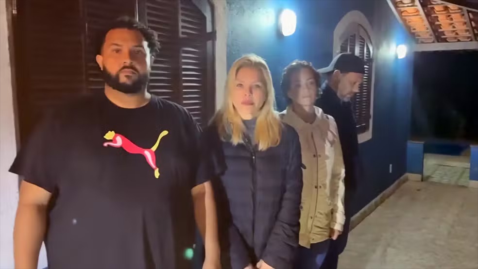 Veronica Costa e os demais condenados após a prisão em Petrópolis, marcando o fim de uma saga judicial de 15 anos. Quatro pessoas, incluindo uma mulher loira, paradas em frente a uma casa azul à noite