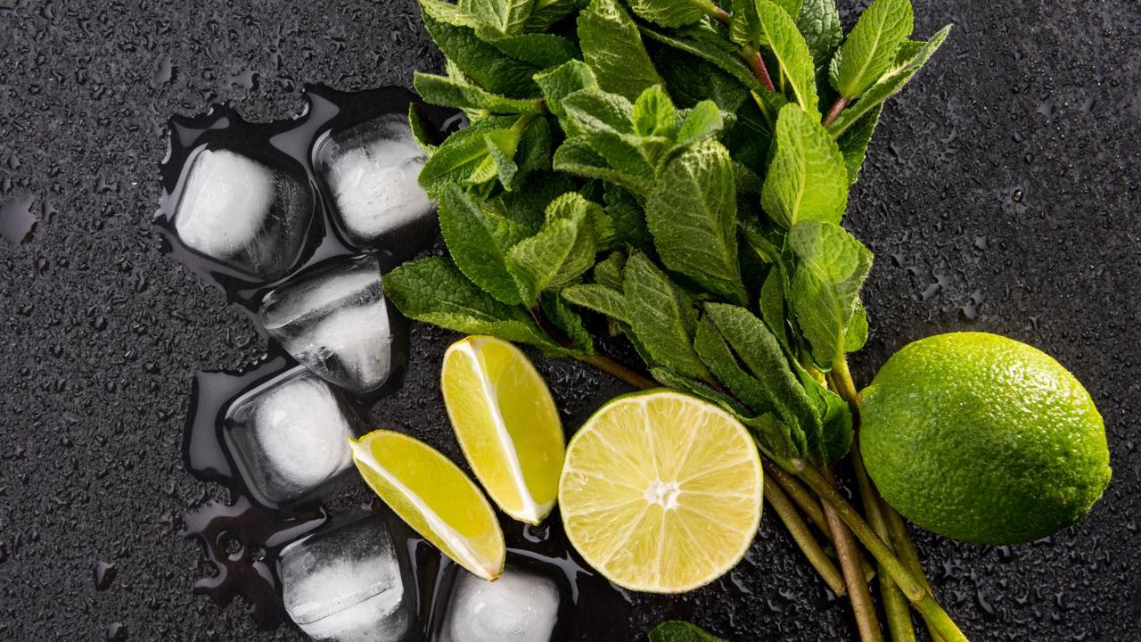Hortelã, limão e gelo, ingredientes essenciais para o preparo de um mojito refrescante, como a receita tradicional. Folhas de hortelã, limões cortados, um limão inteiro e cubos de gelo sobre superfície escura