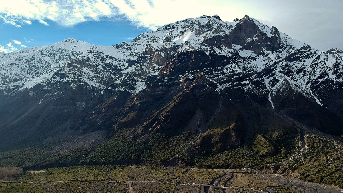 Grande montanha nevada e rochosa sobre vale verde com estradas e céu azul claro.