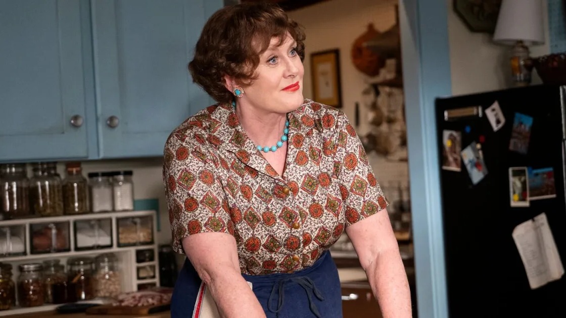 Atriz de cabelo cacheado e camisa estampada com joias em cozinha azul.