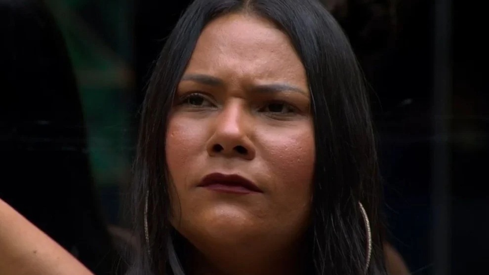 Chaiany, participante do BBB 26, em um momento de reflexão no confinamento. Close-up de Chaiany com cabelo escuro, expressão séria, batom escuro e brincos de argola.