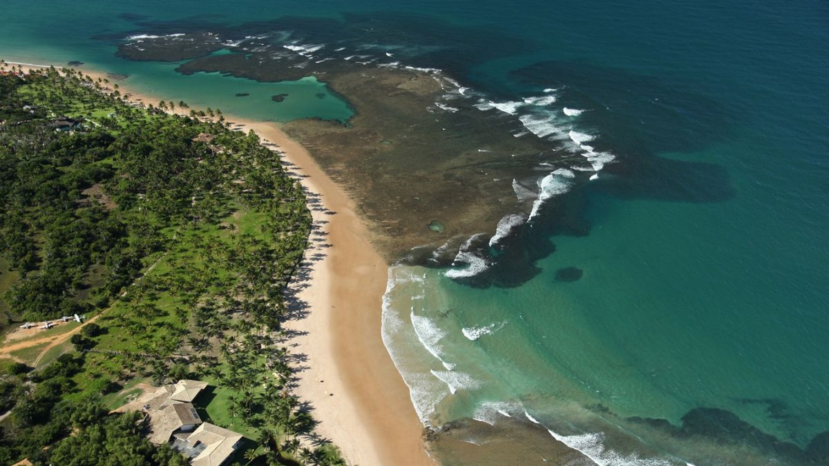 Vista aérea de uma praia tropical com densa vegetação verde, areia clara, mar azul-turquesa e formações rochosas.
