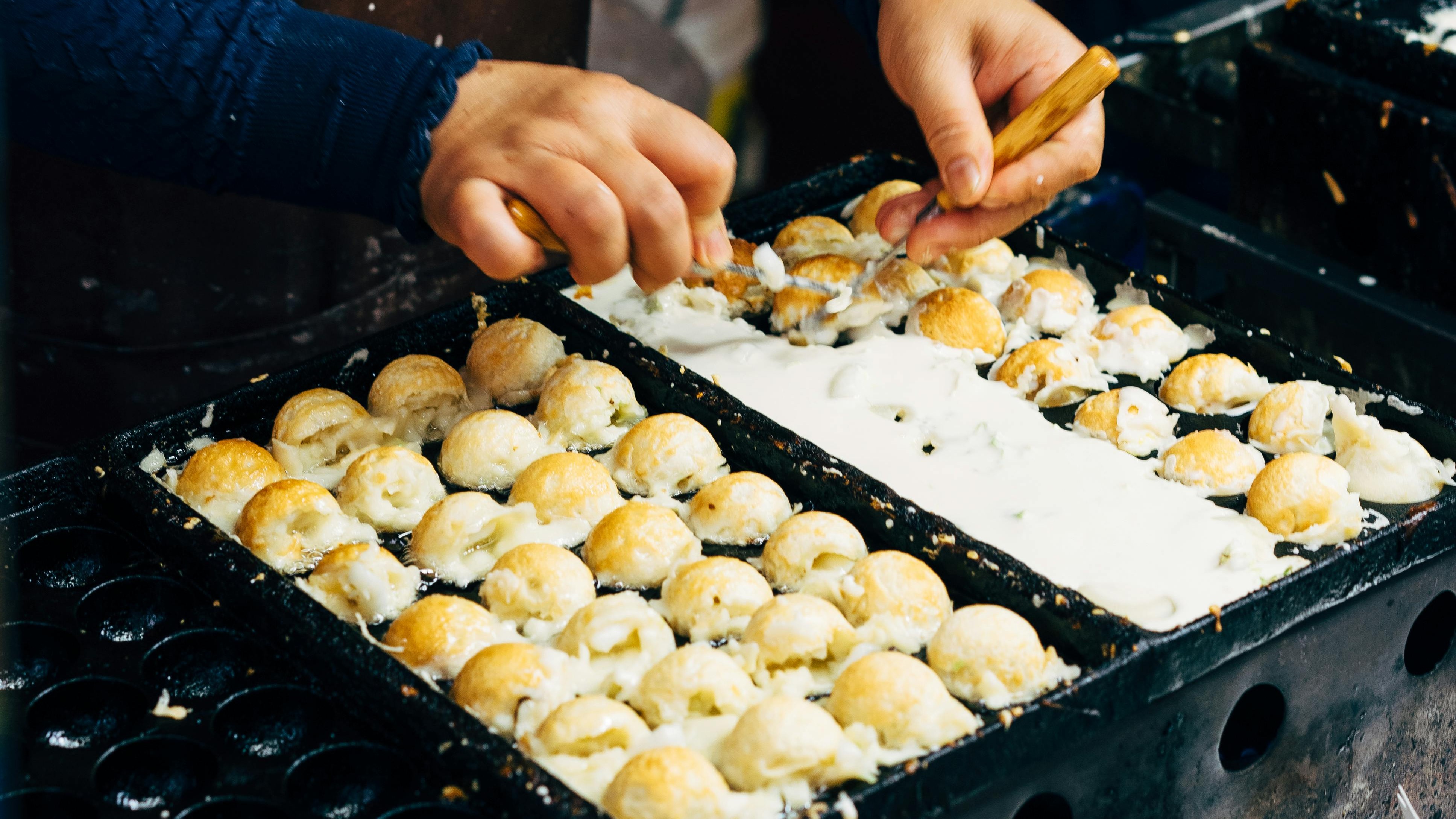 Mãos virando bolinhos de takoyaki em chapa quente com recheio e massa.