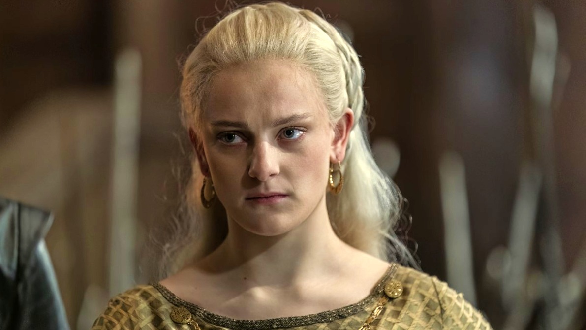 A personagem Helaena Targaryen, cujo destino impacta diretamente a trajetória de Rhaenyra na terceira temporada de A Casa do Dragão. Close-up de mulher loira com vestido dourado e brincos, expressão séria