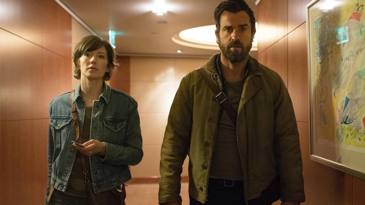 A aclamada série "The Leftovers" explora o luto e a busca por sentido após um evento inexplicável, destacada na lista de sugestões. Mulher de jaqueta jeans e homem barbudo de jaqueta verde caminham por corredor de madeira