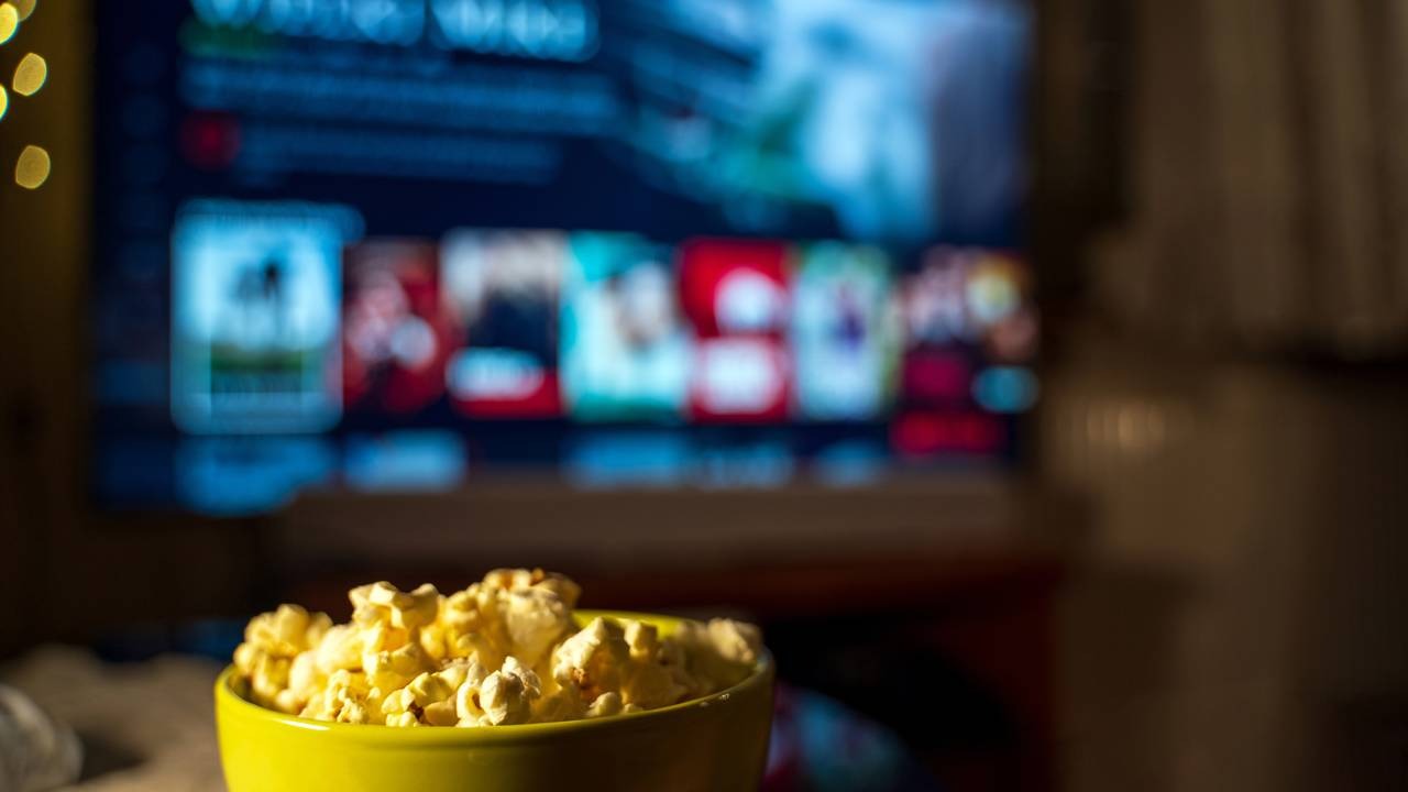 Com a pipoca garantida, explore os melhores romances da Netflix para uma noite especial de Dia dos Namorados. Tigela de pipoca amarela em destaque com tela da Netflix desfocada ao fundo.