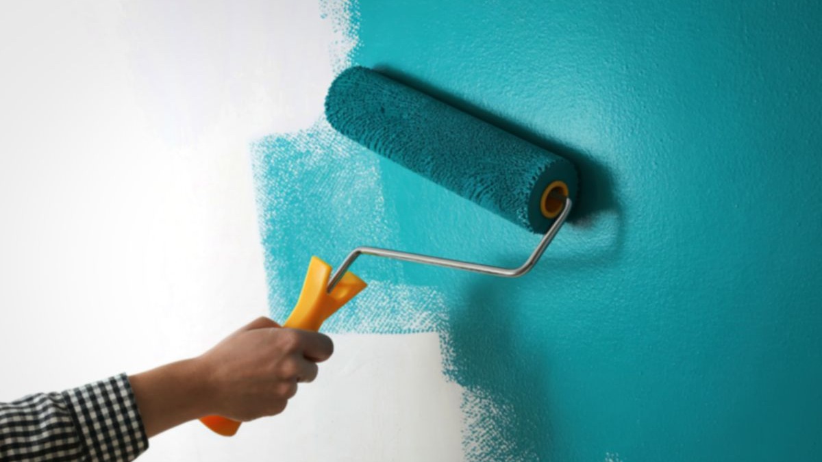 M&atilde;o usando rolo de pintura turquesa para pintar parede branca.