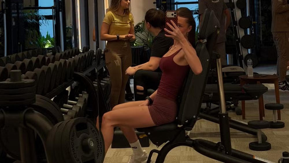 Duda Reis pratica atividade física em academia, hábito fundamental para sua saúde mental e rotina ativa. Mulher sentada em aparelho de academia, tirando selfie com outras pessoas e pesos ao fundo