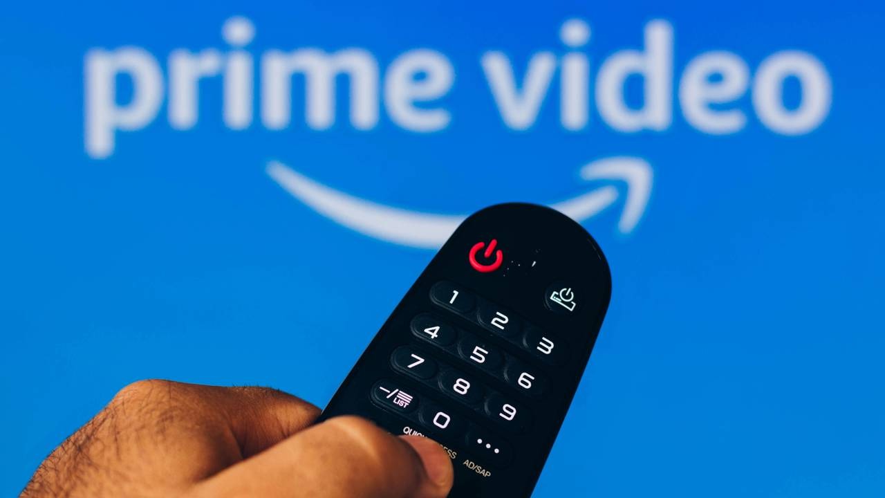 A produção mexicana de true crime "Quem Matou?" está no catálogo do Amazon Prime Video. Mão segura controle remoto preto em frente ao logo Prime Video em tela azul.