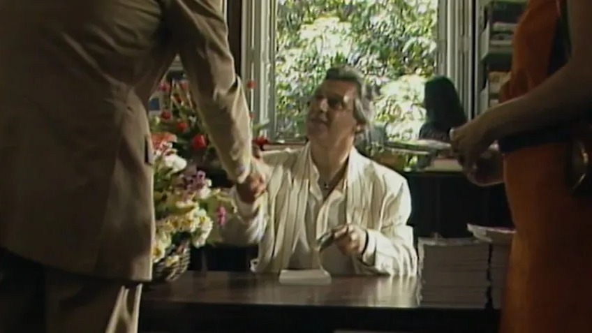 Antonio Fagundes, sentado, aperta a mão de pessoa em terno, com buquê de flores na mesa.