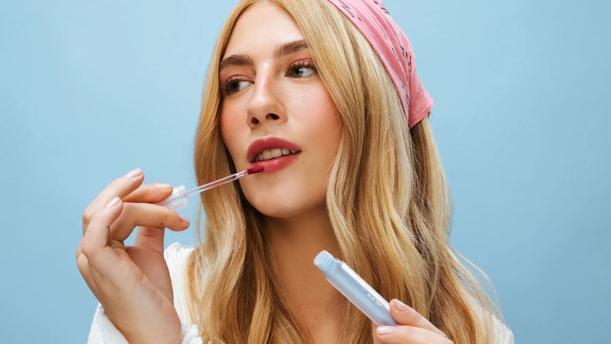 Mulher loira aplicando lip tint nos l&aacute;bios, segurando frasco azul escrito BLUR TINT
