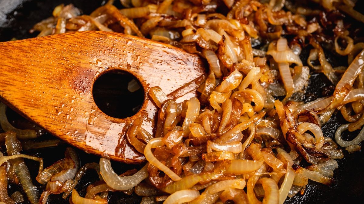 Cebolas caramelizadas douradas em panela preta, sendo mexidas com colher de madeira.