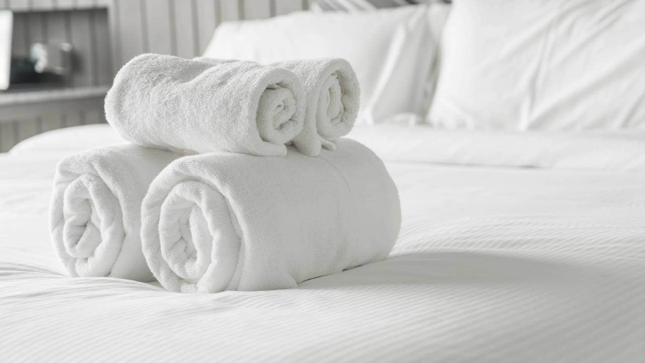 A perfeição hoteleira na arrumação da cama serve como inspiração para começar o dia com produtividade e bem-estar. Quatro toalhas brancas enroladas sobre uma cama de hotel limpa e arrumada