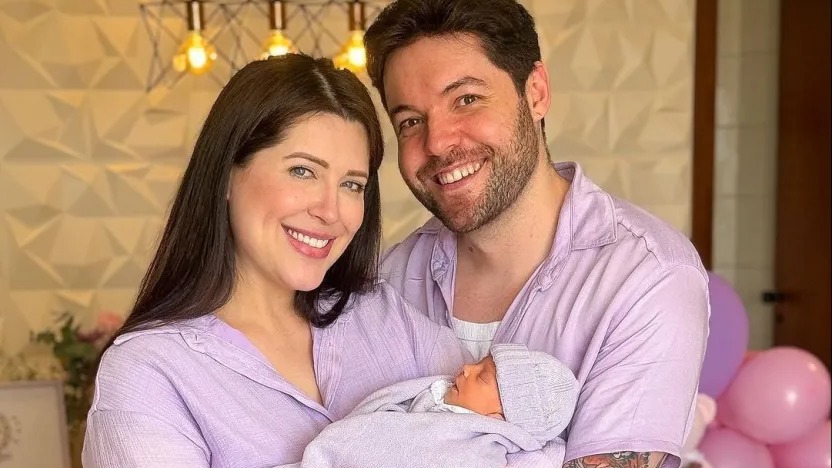 O casal é um dos que formaram família após o reality show. Casal sorridente, segura bebê recém-nascido envolto em manta lilás.