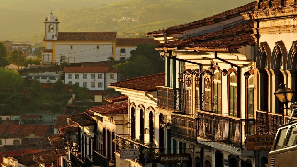 Ouro Preto, Patrimônio Mundial da Unesco, convida à exploração de sua arquitetura colonial e rica história. Vista panorâmica de Ouro Preto com casarões coloniais, igreja e montanhas, sob luz dourada.