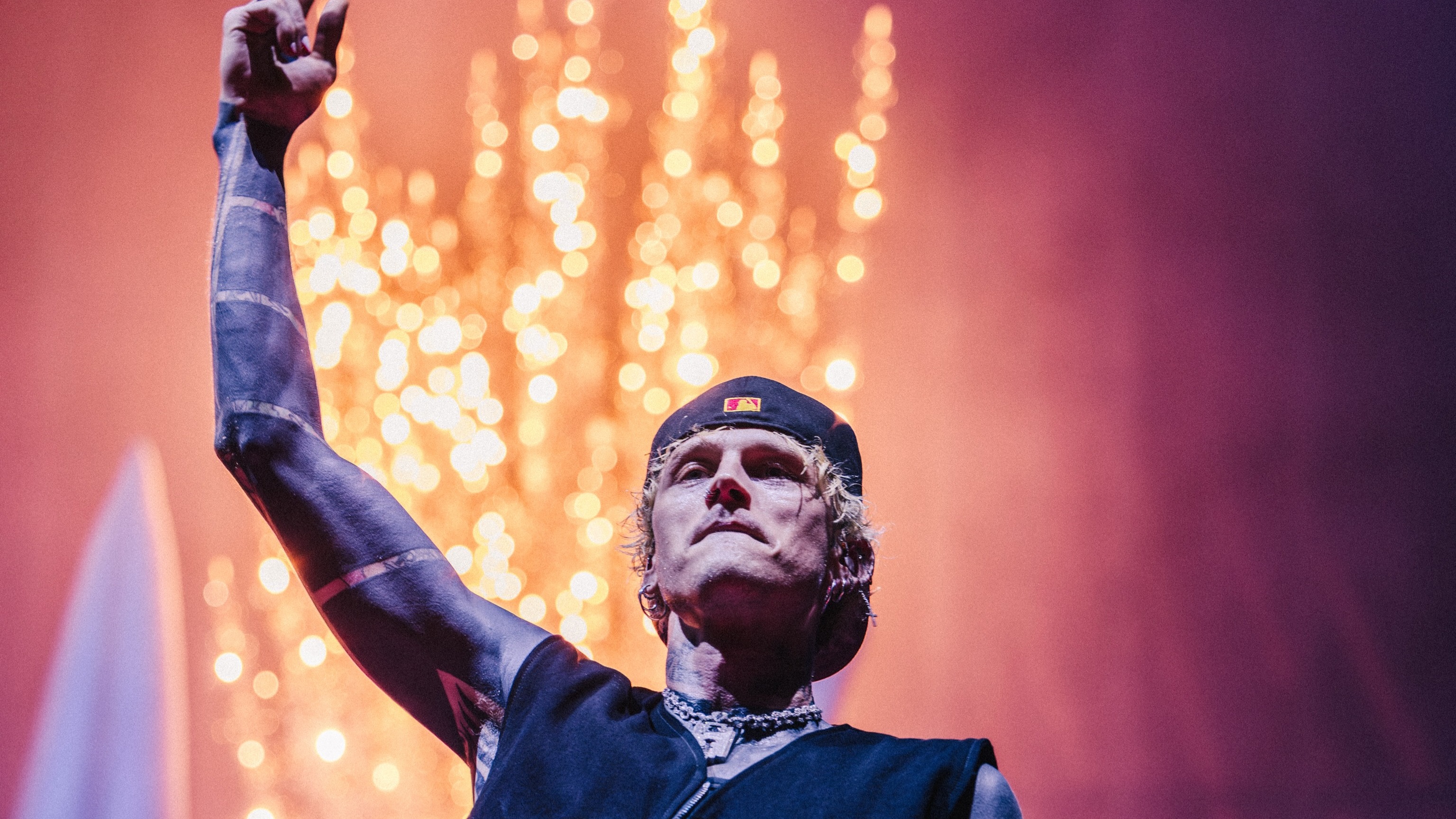 Machine Gun Kelly, que se apresenta no Palco Mundo, é uma das atrações do segundo dia de Rock in Rio 2026 dedicado ao metal. Homem loiro com boné preto, braço tatuado levantado, em palco com luzes bokeh alaranjadas