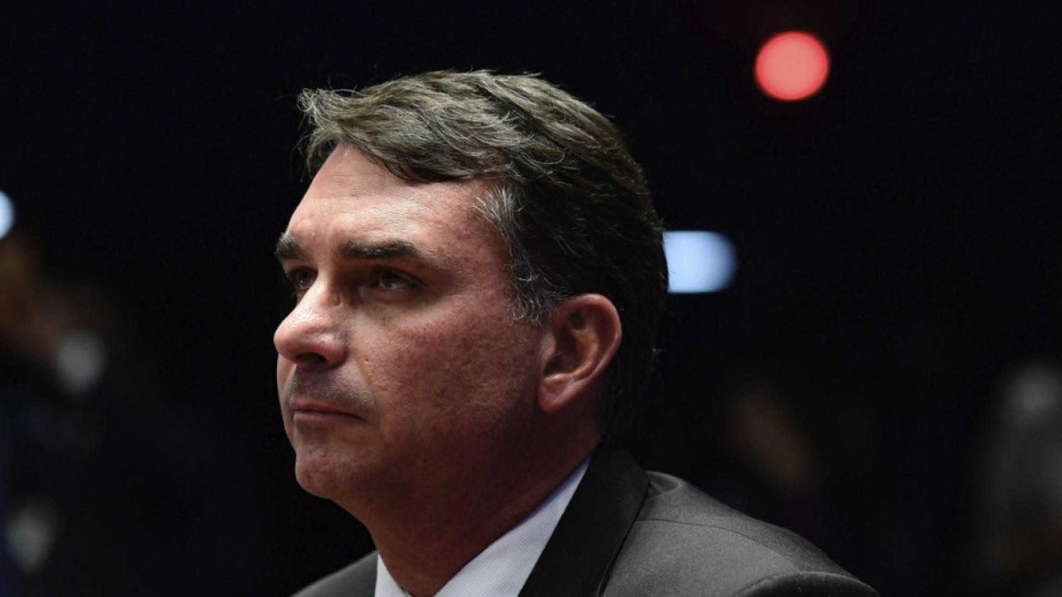 Fl&aacute;vio Bolsonaro, de perfil, com semblante s&eacute;rio e fundo escuro desfocado