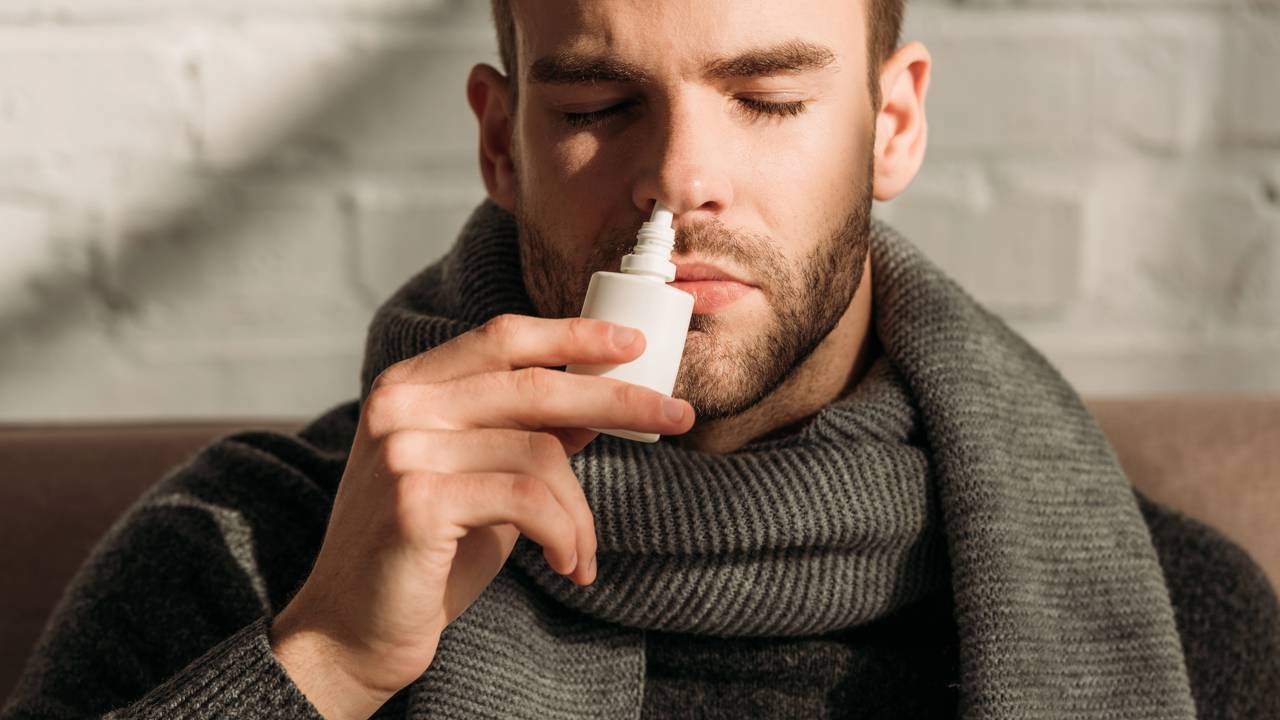 Aliviar a rinite no tempo seco é possível com medidas que combatem a congestão e melhoram a respiração. Homem barbudo com cachecol cinza aplicando spray nasal branco no nariz com olhos fechados