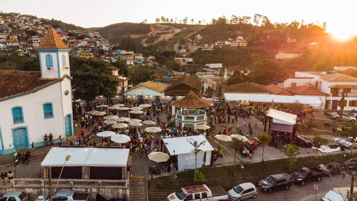 Pra&ccedil;a movimentada em cidade mineira, com igreja branca, coreto, barracas, pessoas e montanhas ao fundo.
