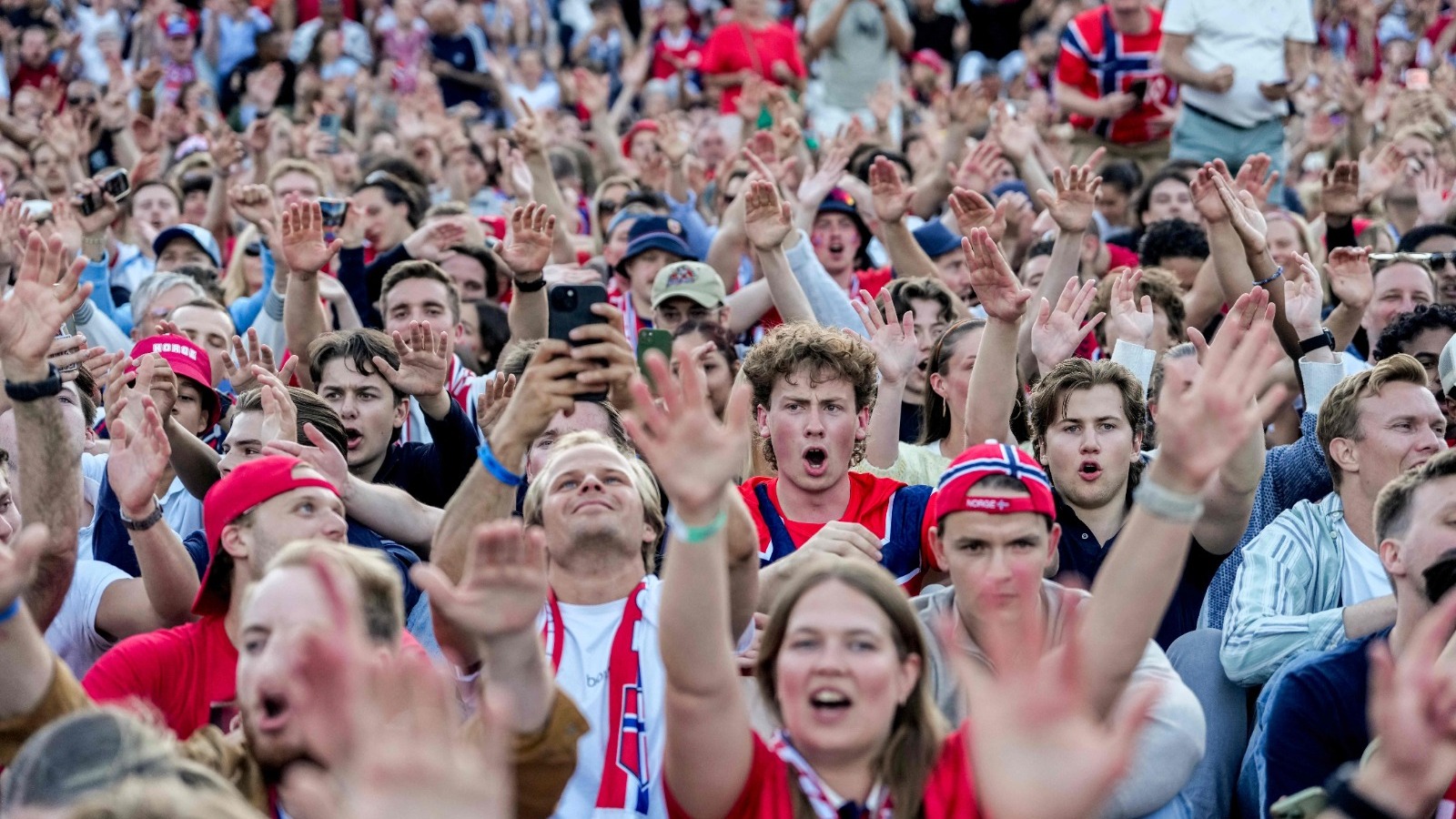 Multid&atilde;o de torcedores noruegueses com as m&atilde;os para cima, alguns usando bon&eacute;s com a bandeira da Noruega.