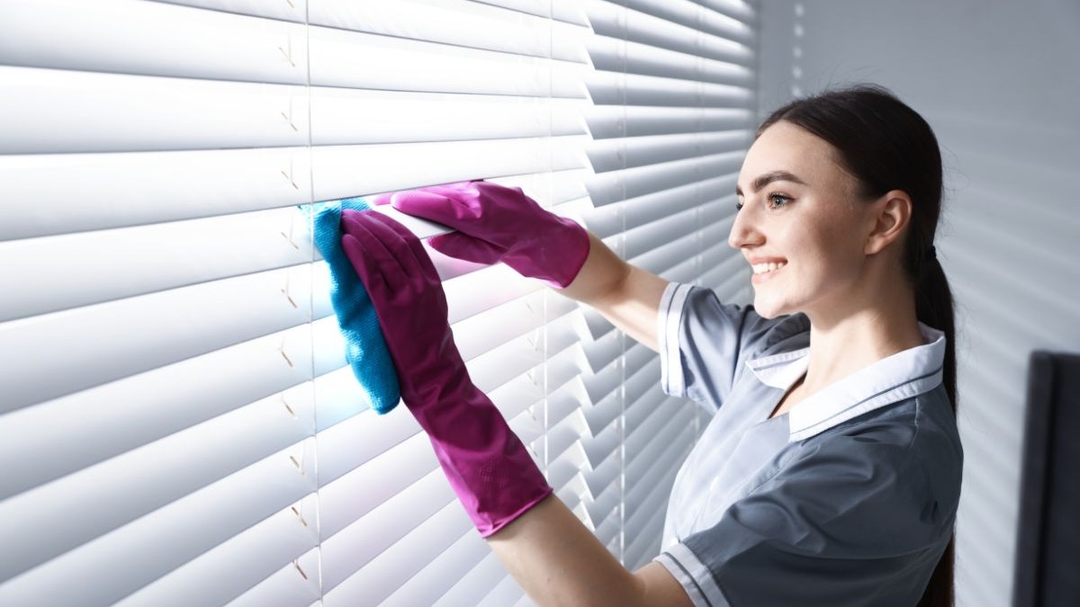 Mulher sorrindo, usando luvas rosa, limpando persianas brancas com pano azul.