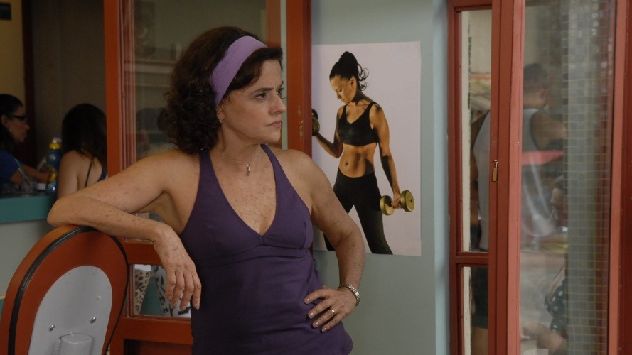 Mulher de meia-idade, em top roxo e faixa na cabe&ccedil;a, com olhar pensativo, encostada em cadeira.